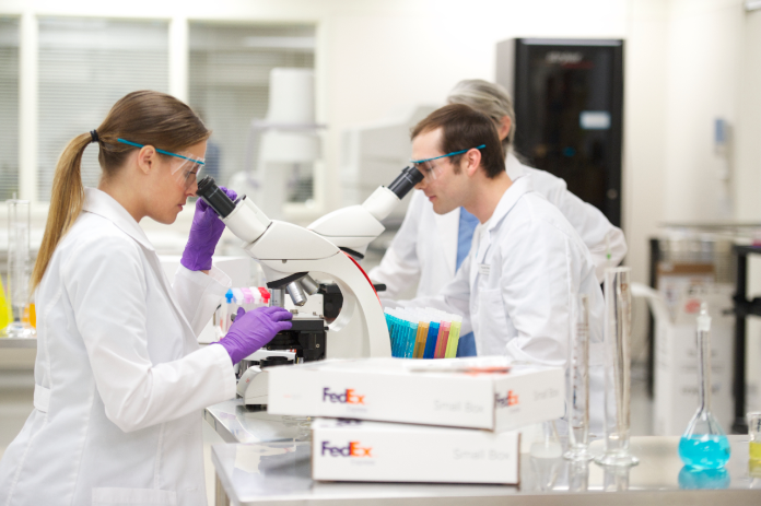 Two lab techs use a high-power microscope to view a slide