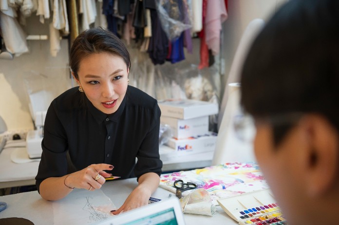 A clothing designer surrounded by fabric and patterns chats with her colleague.