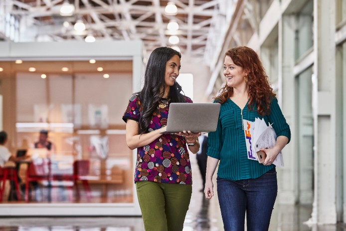 Two women walking and talking while looking at a laptop