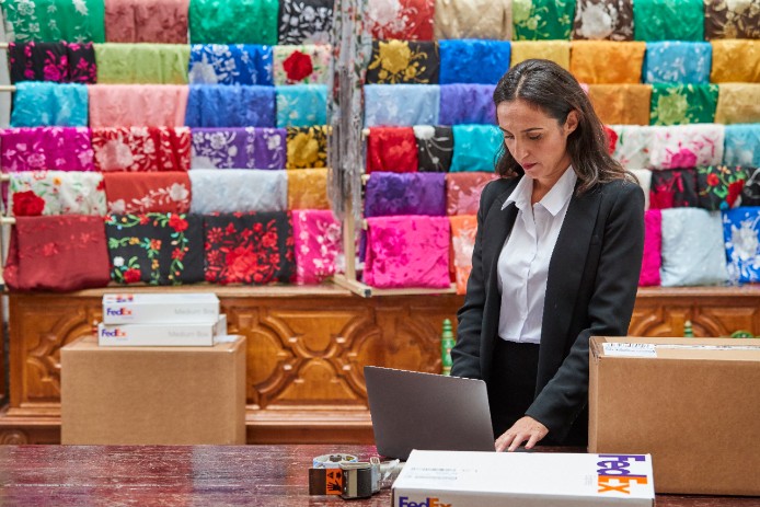 Woman standing in front of shelves of colorful fabric looking at her laptop on a desk next to a FedEx box