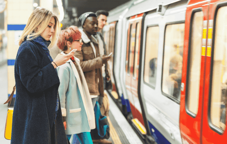 Wartende Menschen am Bahnhof neben einer U-Bahn, eine Person blickt auf ihr Telefon