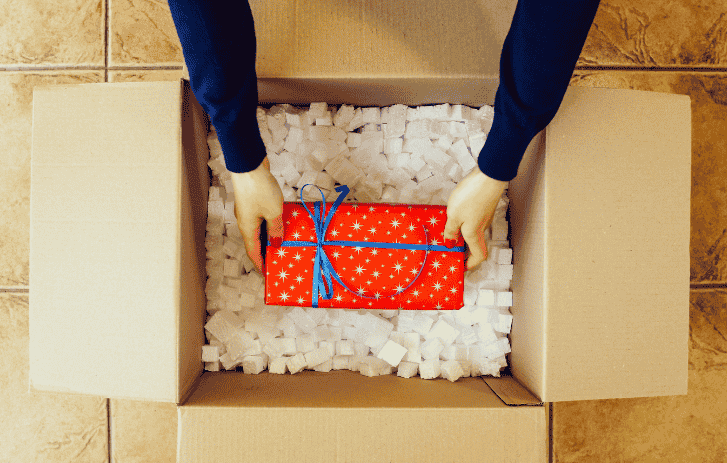Someone reaching into an open cardboard box filled with packing material, adjusting a red gift box tied with a blue ribbon. 