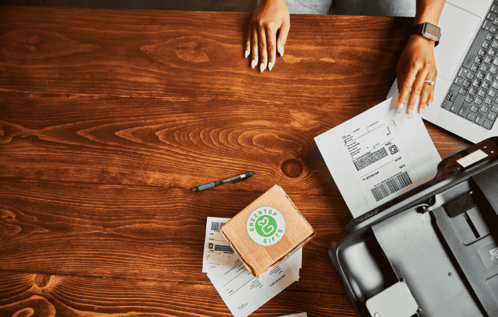 An eco-friendly package sits on a table while someone prints a shipping label nearby. 