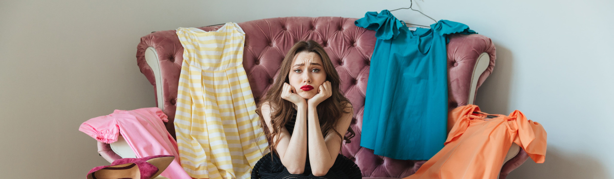 A woman sits on a couch surrounded by clothes and shoe boxes, looking indecisive. 