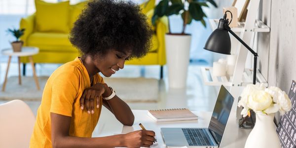 Woman writing in a notebook at a desk with a laptop, papers, a desk lamp, and flowers. 