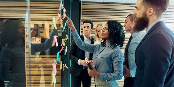 A group of people gathered around a glass wall with colorful sticky notes as a woman in a blue shirt points and leads the discussion.