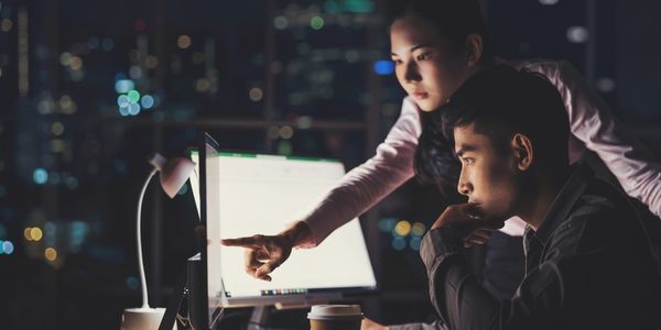 Two colleagues work late at a desk as one points to a bright computer screen, with a coffee cup, desk lamp alongisde them.