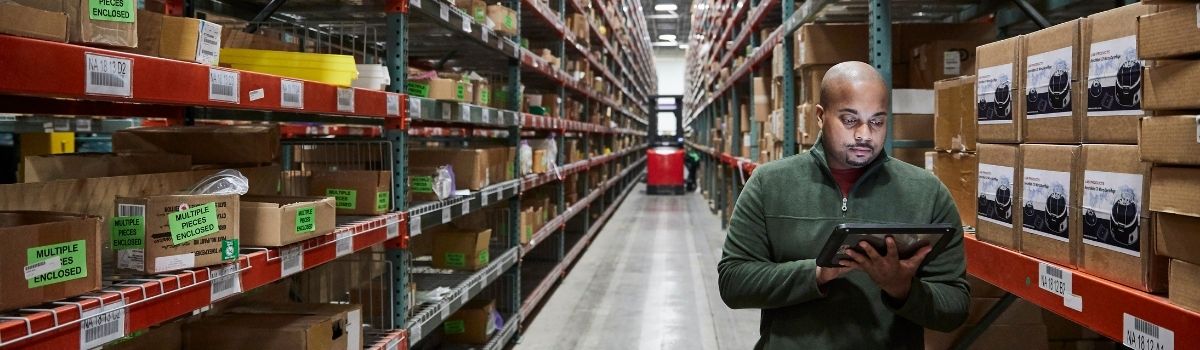 A man stands in a long warehouse aisle, looking at a tablet while shelves are stacked with boxes and pallets in the background. 