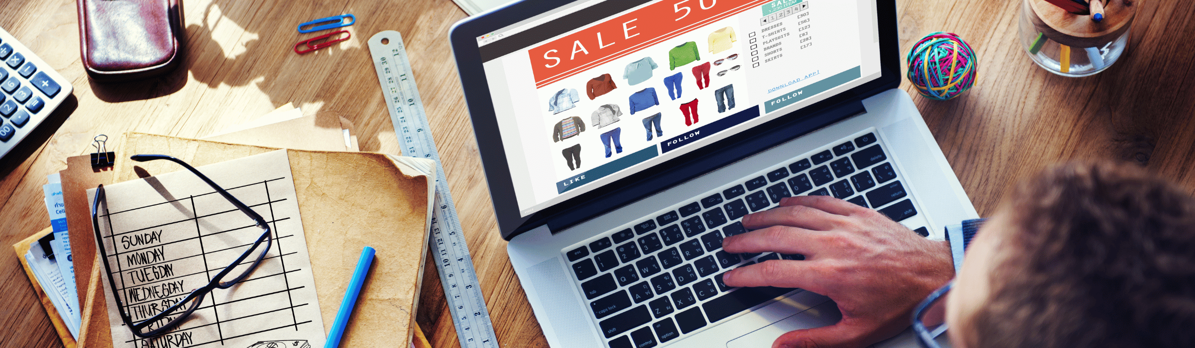 Mężczyzna szukający na komputerze ubrań w promocyjnych cenach, otoczony porozrzucanymi artykułami biurowymi.