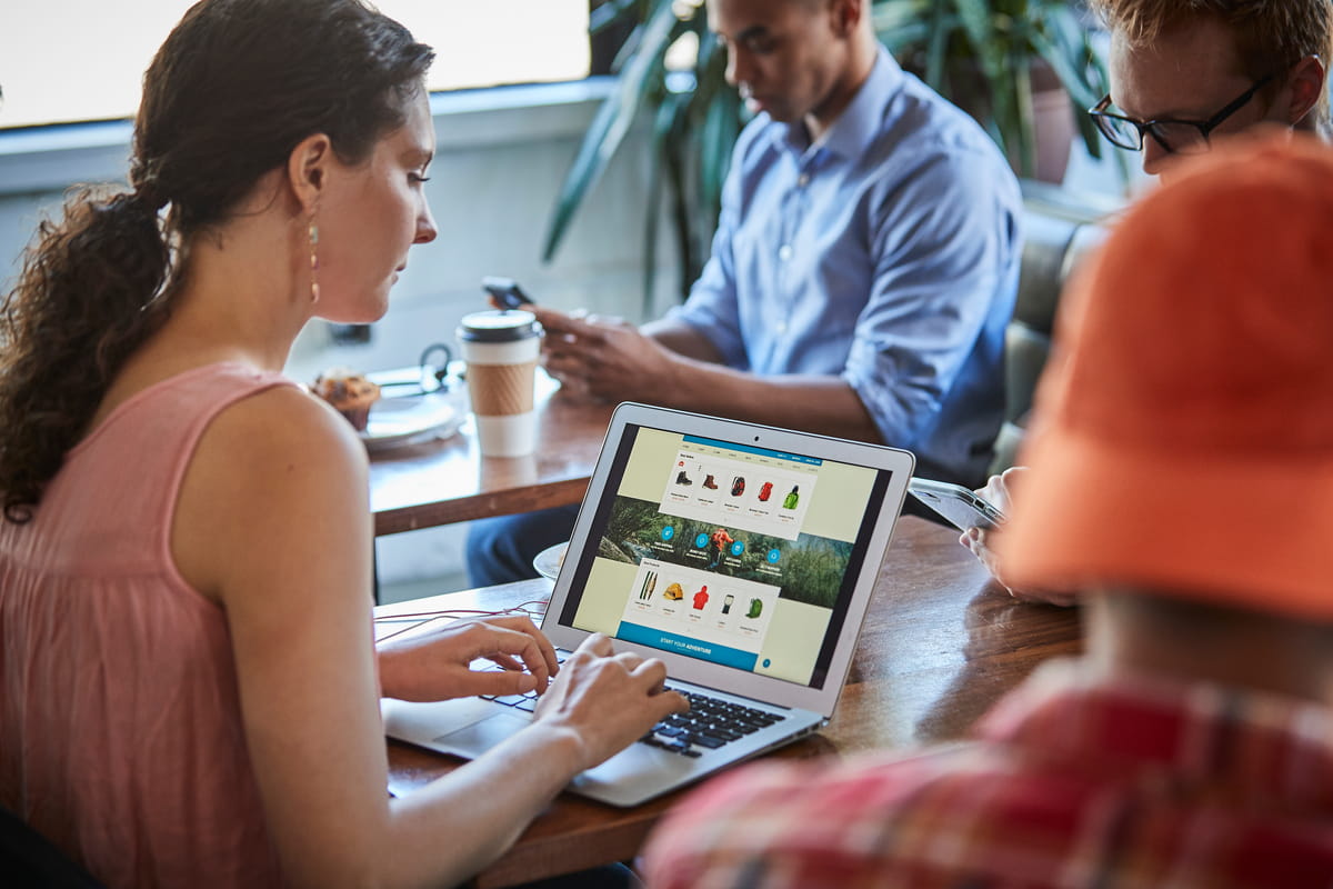 Une femme assise à la table d’un café, réalisant des achats en ligne sur son ordinateur portable dans un environnement lumineux et détendu. 
