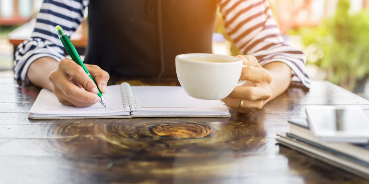 A person sitting in front of a wooden table, writing in an open spiral notebook with a green pen while holding a white mug of coffee. 