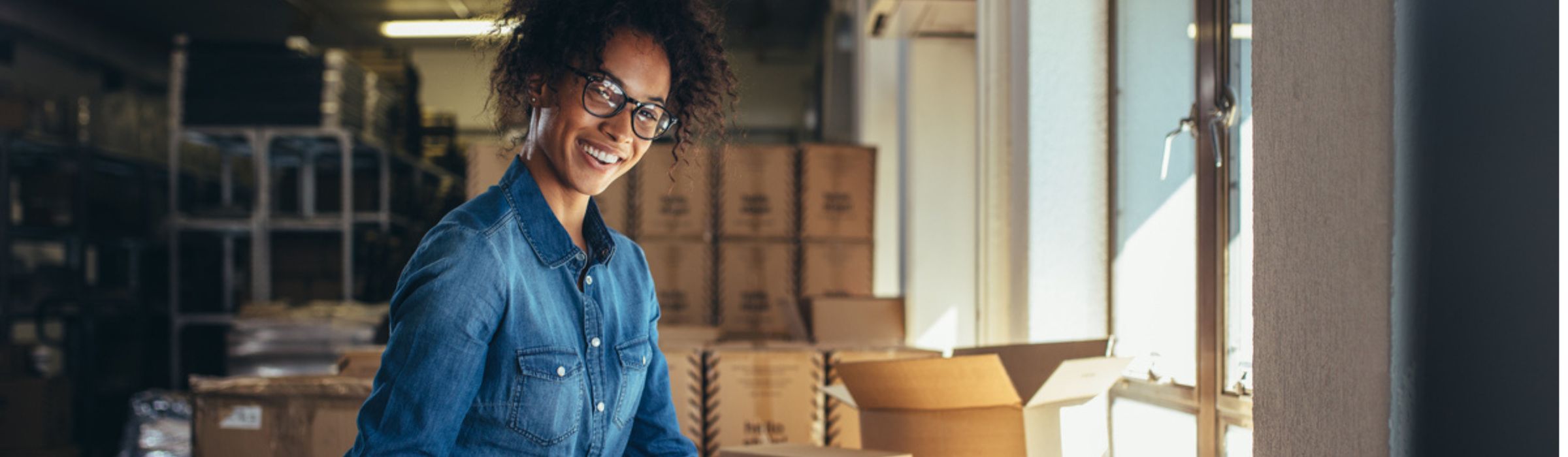 A smiling woman standing in a sunlit warehouse with stacked cardboard boxes and shelves. 