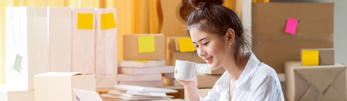 Een vrouw in een wit overhemd zit achter een houten bureau, typt op een laptop en neemt een slokje uit haar mok, omringd door ingepakte dozen en verzendmateriaal op de achtergrond.