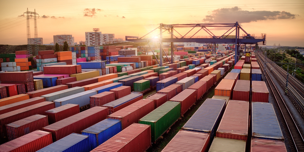 Rows of colorful shipping containers at sunset, a gantry crane overhead and railway tracks to the right. 