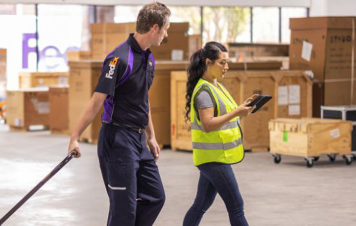 Lagermitarbeiter*innen verwalten Sendungen und Bestände mithilfe eines Hubwagens und eines Tablets inmitten verpackter Waren.