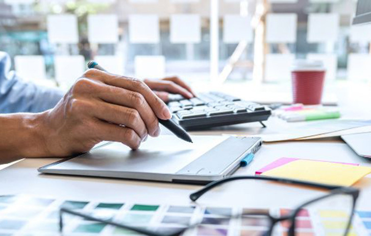 A man using a tablet and computer at a creative workspace with design materials and colour samples on the desk.