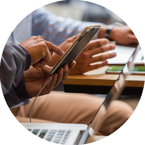 Two people are working side by side at a table, one using a smartphone while the other types on a laptop