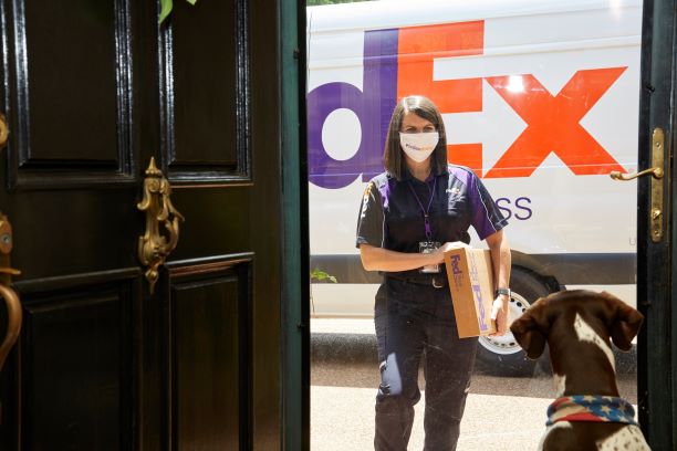 FedEx courier delivers a package to a house while a dog waits at the door