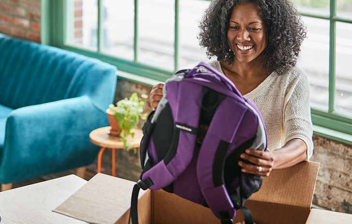A woman holding up a backpack from an open brown shipping box