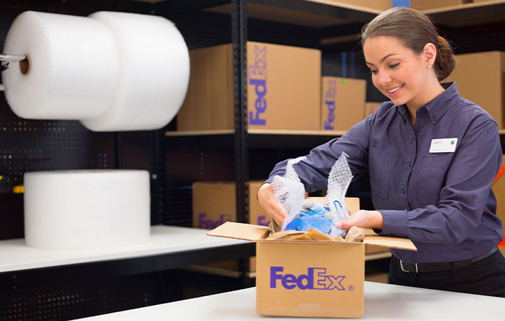 Image of a FedEx employee packing an item wrapped in bubblewrap into a brown FedEx box with packing supplies in the background.