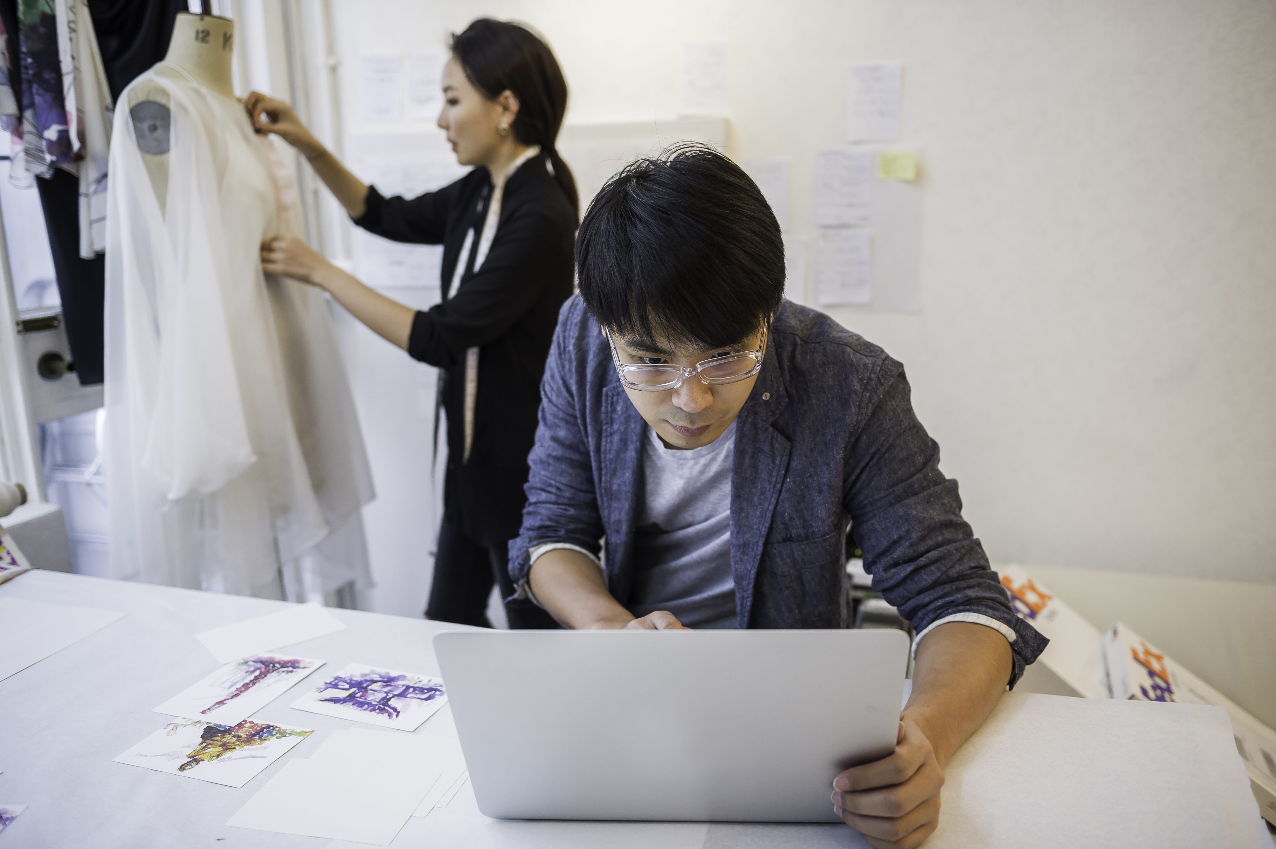 A fashion industry businesswoman alters a gown while a businessman types on his laptop.