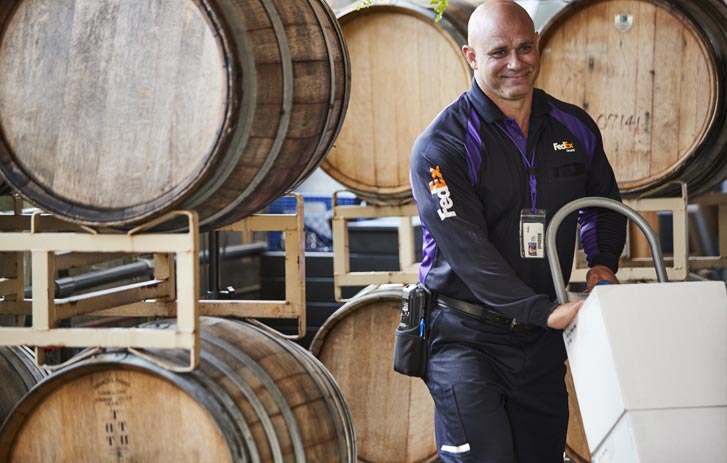 A photo of a FedEx worker pushing boxes on a dolly near wooden wine barrels