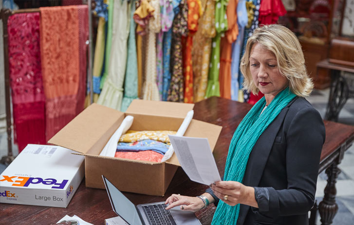 Woman holding papers at a laptop with a FedEx box and a brown box with fabric and clothes are behind her.