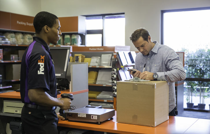 A photo of a man standing at a FedEx Office counter and signing a form to ship a package while a FedEx worker stands by to help