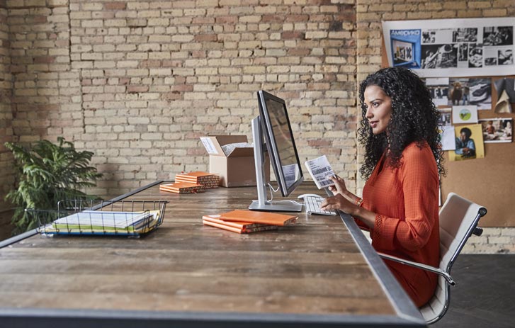 A photo of a woman holding a shipping label while sitting at her desktop computer.