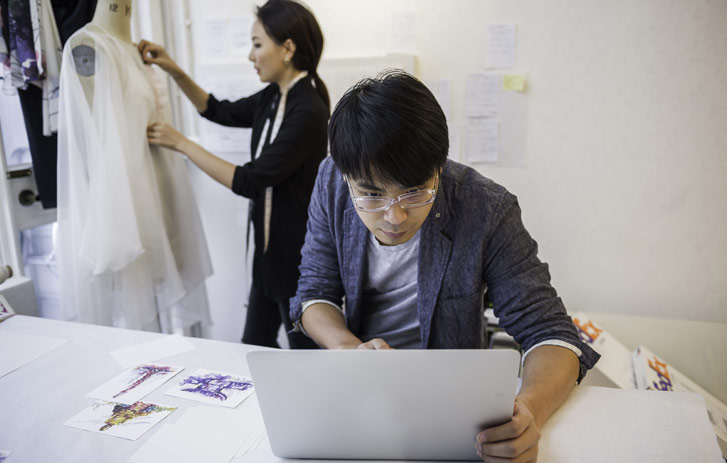 A fashion industry businesswoman alters a gown while a businessman types on his laptop.