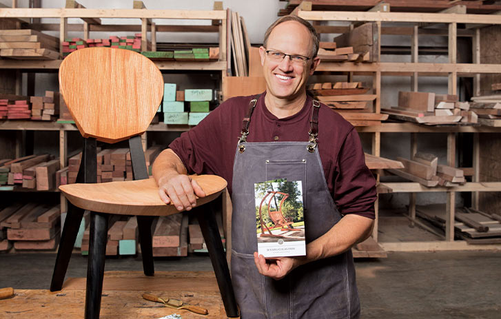 A photo of Brian Boggs of Brian Boggs Chairmakers, Inc. leaning against a chair and holding a brochure in a woodworking shop
