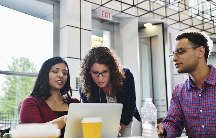 A photo of two women and a man sitting at a table looking at a laptop and having a discussion.