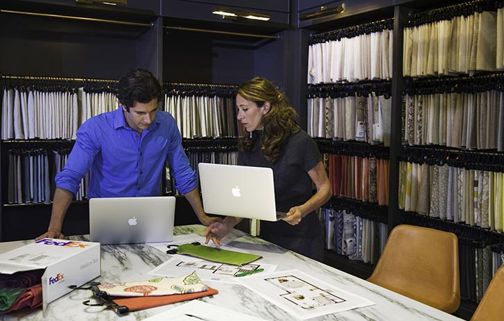 A man and a woman stand looking at two laptops while talking with papers spread out on a table before them.