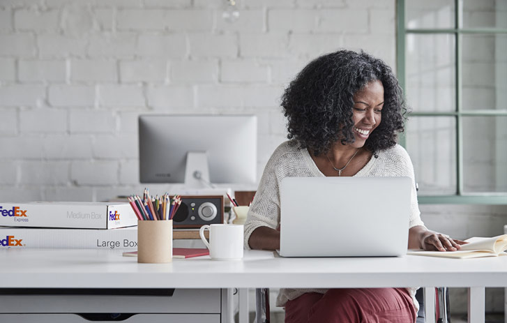 Woman using laptop with FedEx boxes on table.