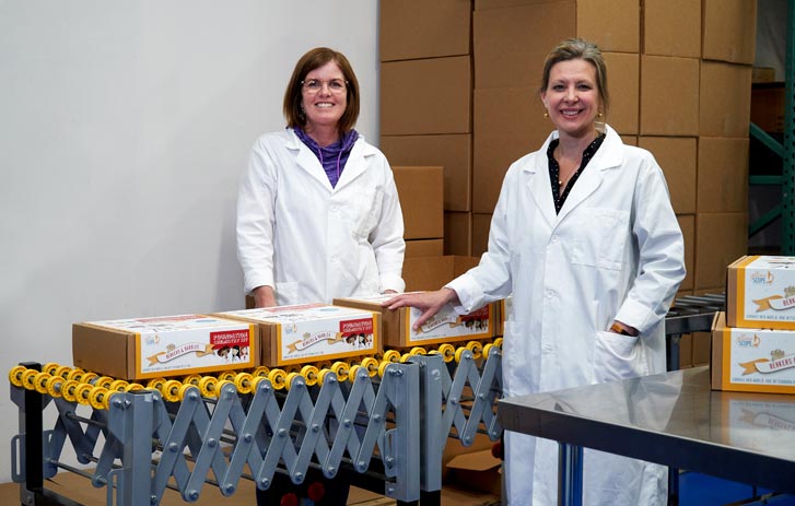 A photo of Kelly McCollum and Marcie Colledge, owners of Yellow Scope, standing with lab coats on in a warehouse next to their chemistry kits on a conveyor belt.