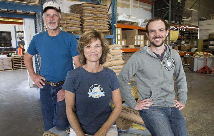 Scratch & Peck A photo of Dennis Meade, Diana Ambauen-Meade, and Byron Meade from Scratch and Peck sitting in a warehouse with feed bags in the background