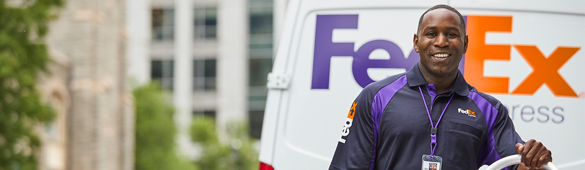 A smiling FedEx courier stands in the foreground of a truck.