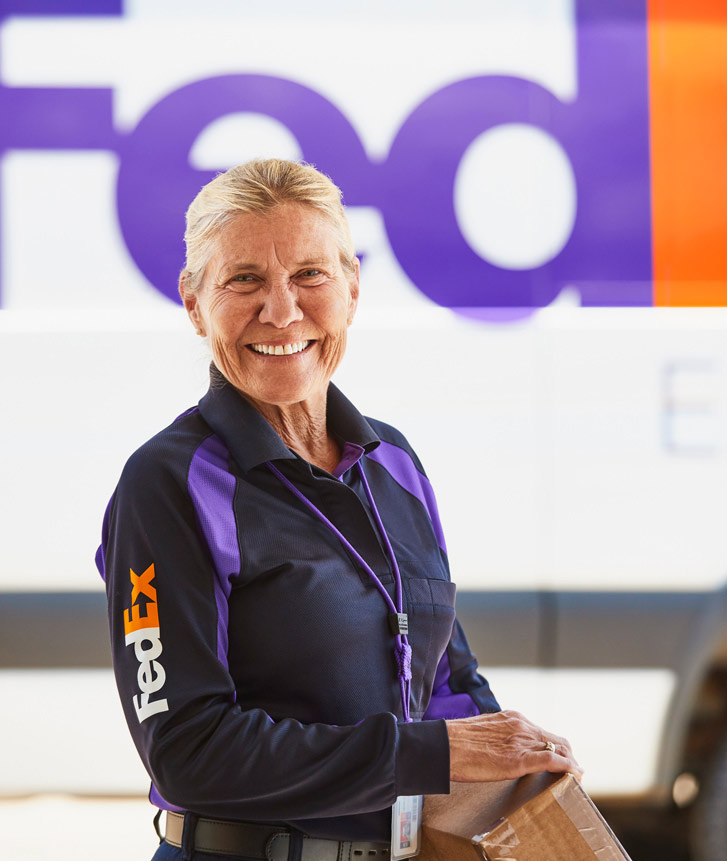 A FedEx courier smiling and holding a brown box in front of a FedEx truck