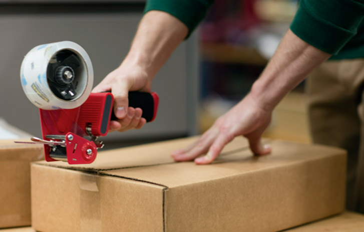 An individual using a tape gun to seal a brown box with clear packing tape.