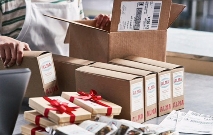 A business owner packing boxed products and boxed products with bows into a brown box with a white shipping label.