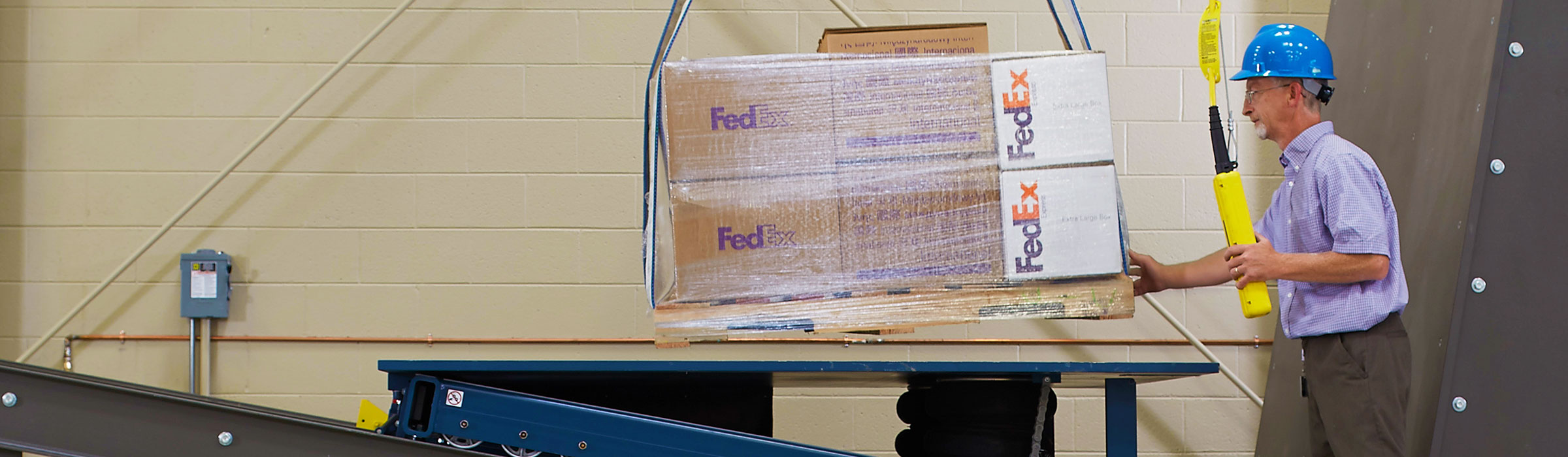 FedEx Packaging Lab employee wearing a hard hat performing a test on multiple boxes wrapped on a pallet at a table.