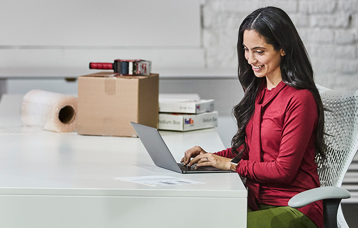 A woman working on a laptop with a brown box, bubble wrap and packaging tape stacked next to her.