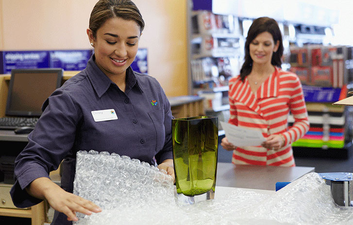FedEx employee wrapping a green, glass vase in bubble wrap, with a customer standing at the counter waiting holding a piece of paper. Packing supplies coupons.