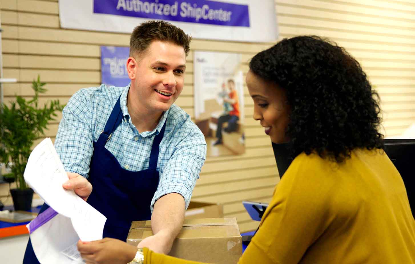 Inside a FedEx Authorized ShipCenter, a smiling employee is showing documents to a smiling customer.